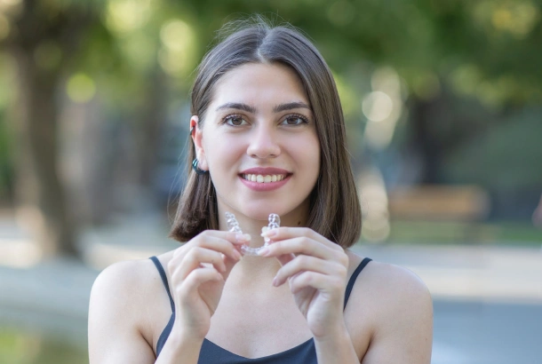 A woman holding a clear aligner and smiling outdoors near Denver Orthodontics and Dental Lounge in Denver CO