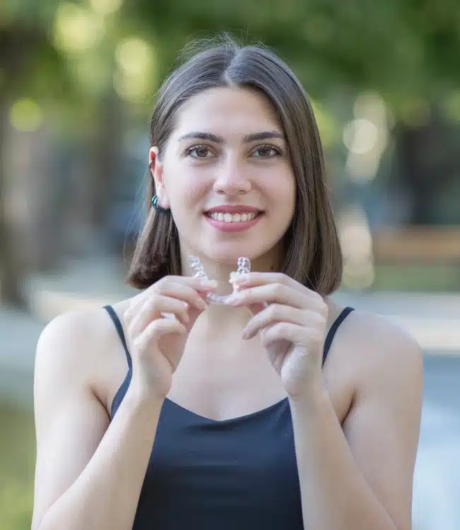 A adult woman smiling while holding Invisalign clear aligners outdoors