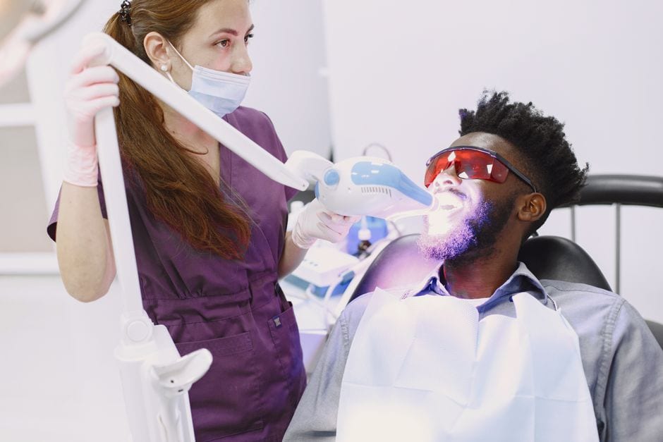 Dentist performing teeth whitening procedure on patient with specialized equipment.