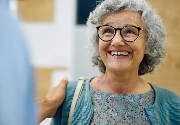 Smiling older woman with gray hair and glasses wearing a teal cardigan in a dental office setting.