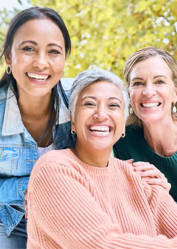 Three smiling women outdoors, representing confidence, healthy smiles, and positive well-being.