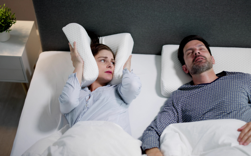 Woman covering her ears with a pillow while her partner snores from sleep apnea in bed