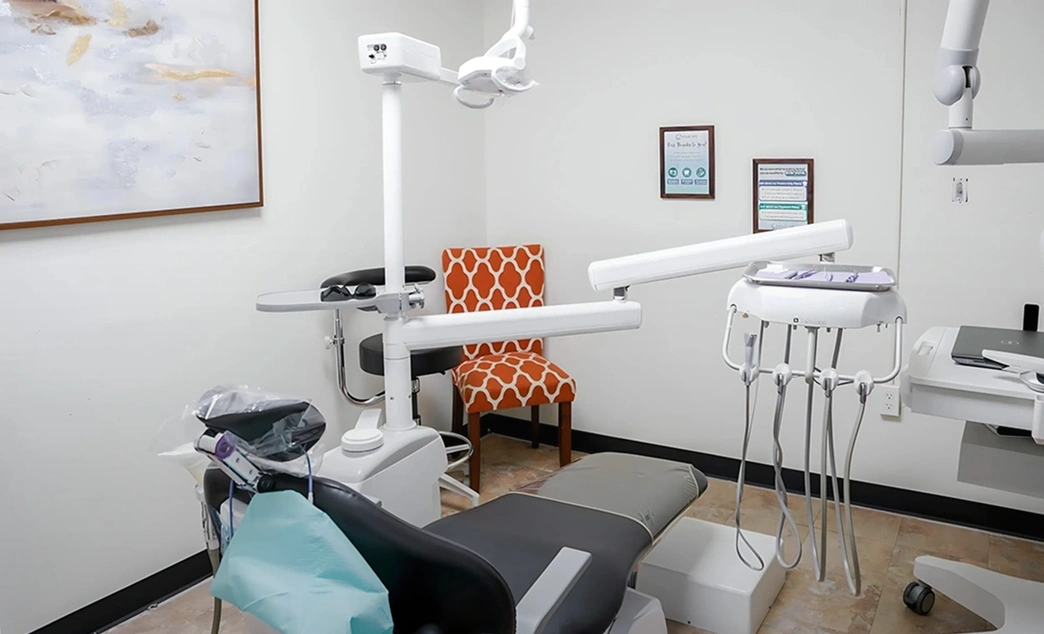 Modern dental operatory room with a dental chair, overhead light, instruments, and an orange patterned patient chair in the corner.
