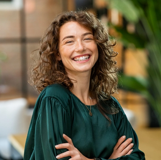 A smiling young woman with curly brown hair wearing a dark green top, standing with arms crossed and showcasing a bright, confident smile.