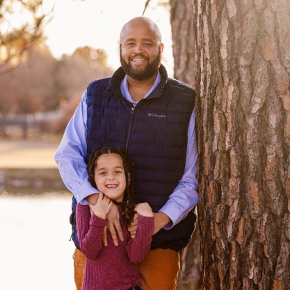 Dr. Talon Davis smiling next to a pine tree outdoors with his young daughter standing in front of him.