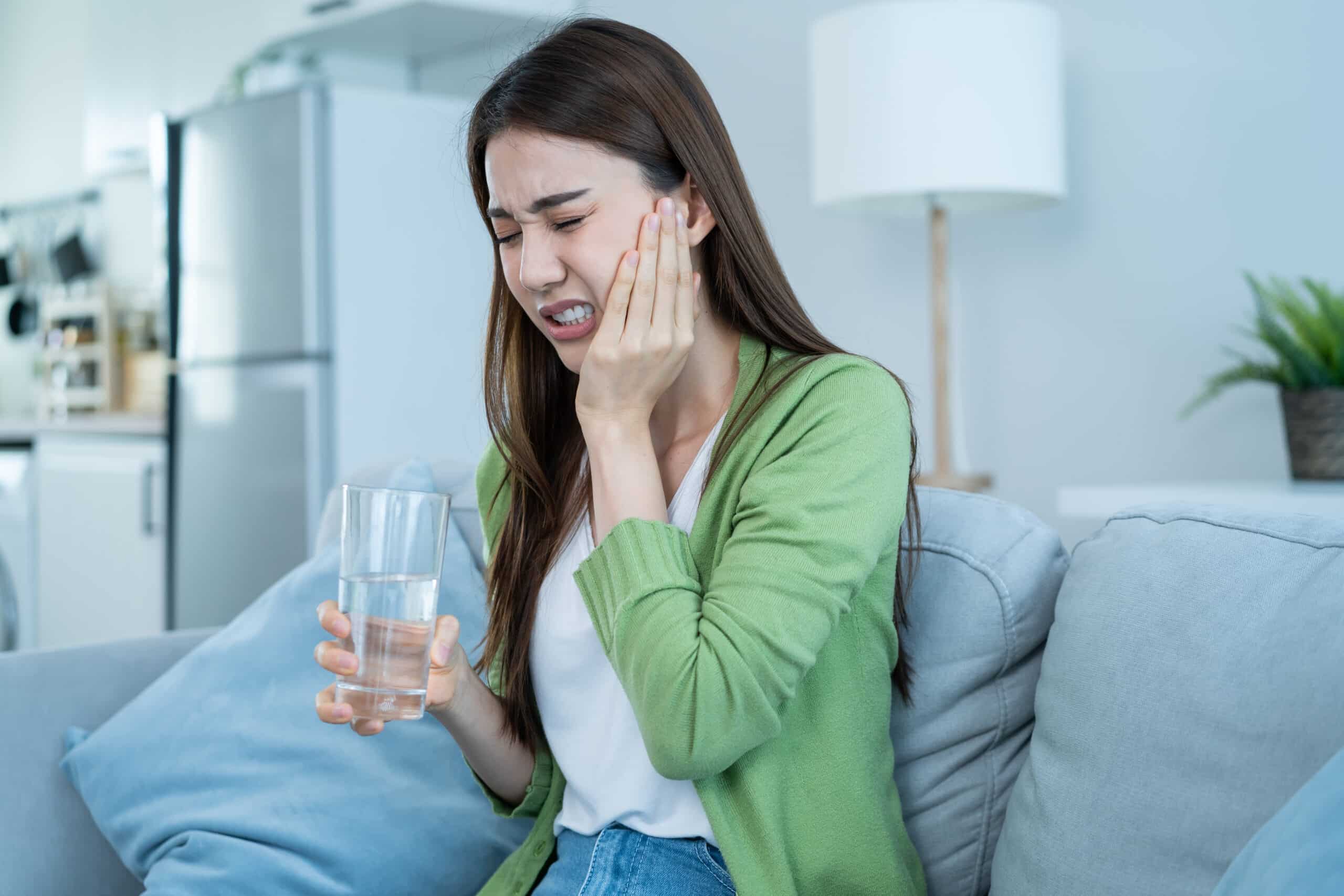 Asian beautiful woman feel terrible toothache after drink cold water. Attractive female sit on sofa in living room touching cheek, feel hurt and suffering from sensitive tooth ache, pain and cavities.