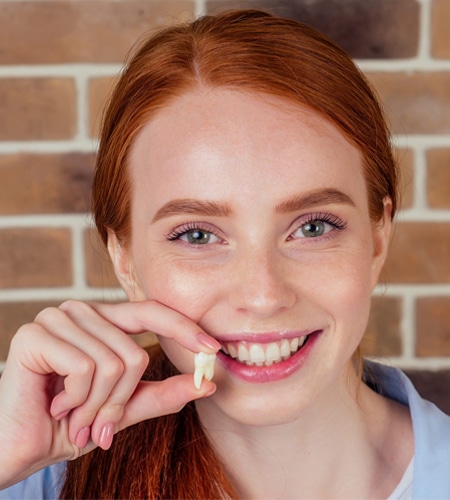 Young woman smiling and holding a tooth in front of a brick wall after a successful tooth extraction in Charlotte NC