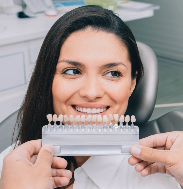 Woman in dental chair smiling as dentist holds a veneer shade guide for custom dental veneers