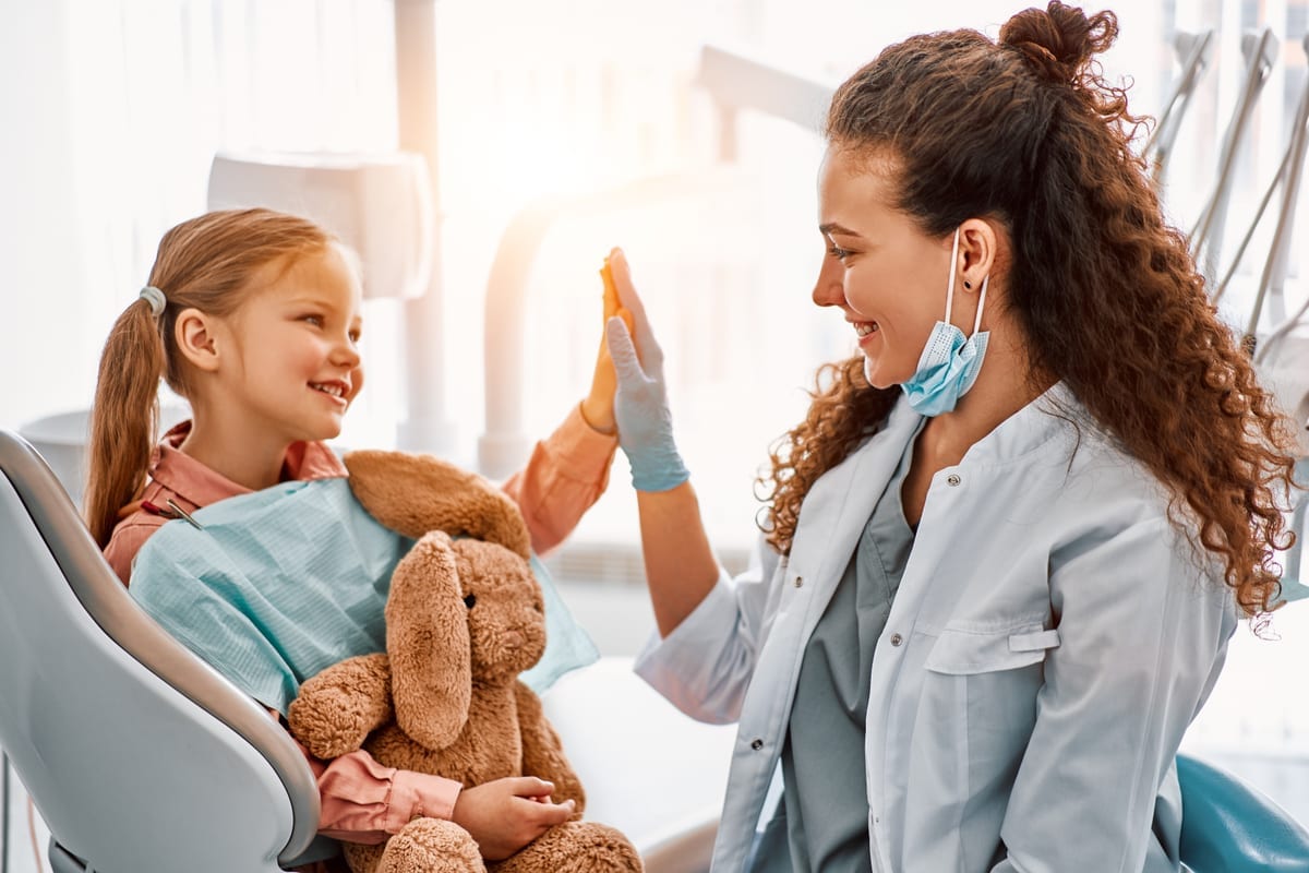 Pediatric dentist giving high-five to smiling child patient holding teddy bear in dental office.