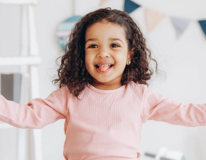Happy young girl with curly dark hair smiling with arms outstretched in bright dental office.